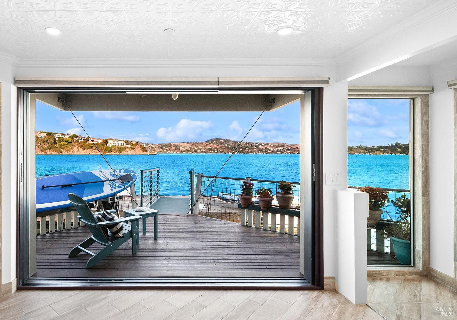 Wall of disappearing glass doors fully retracted, framing the wraparound deck, paddleboard, and panoramic view of Tiburon and the bay from the great room.