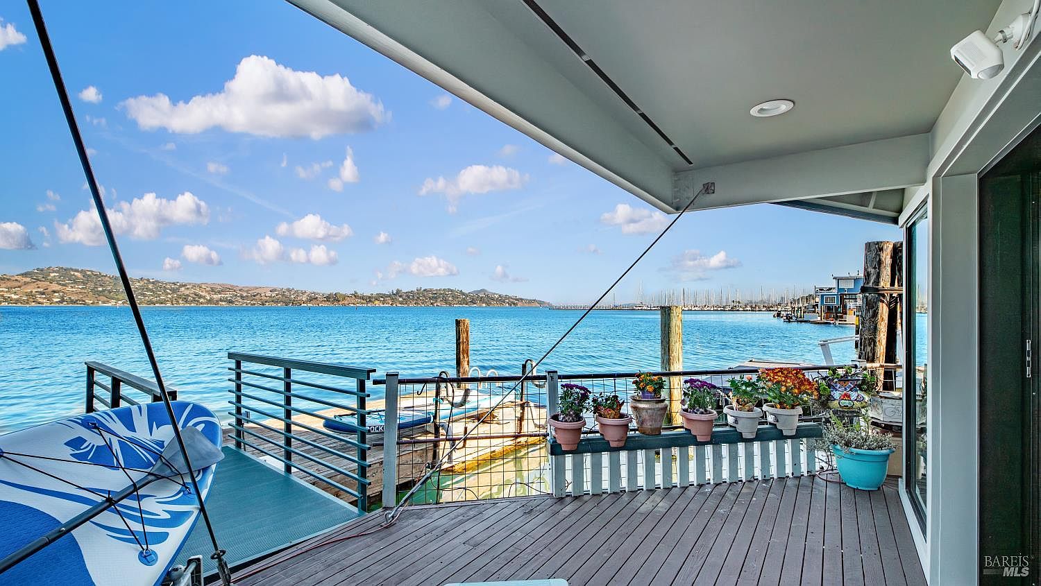Wraparound deck with weathered planking, potted flowers along the rail, and the open bay stretching to the marina.