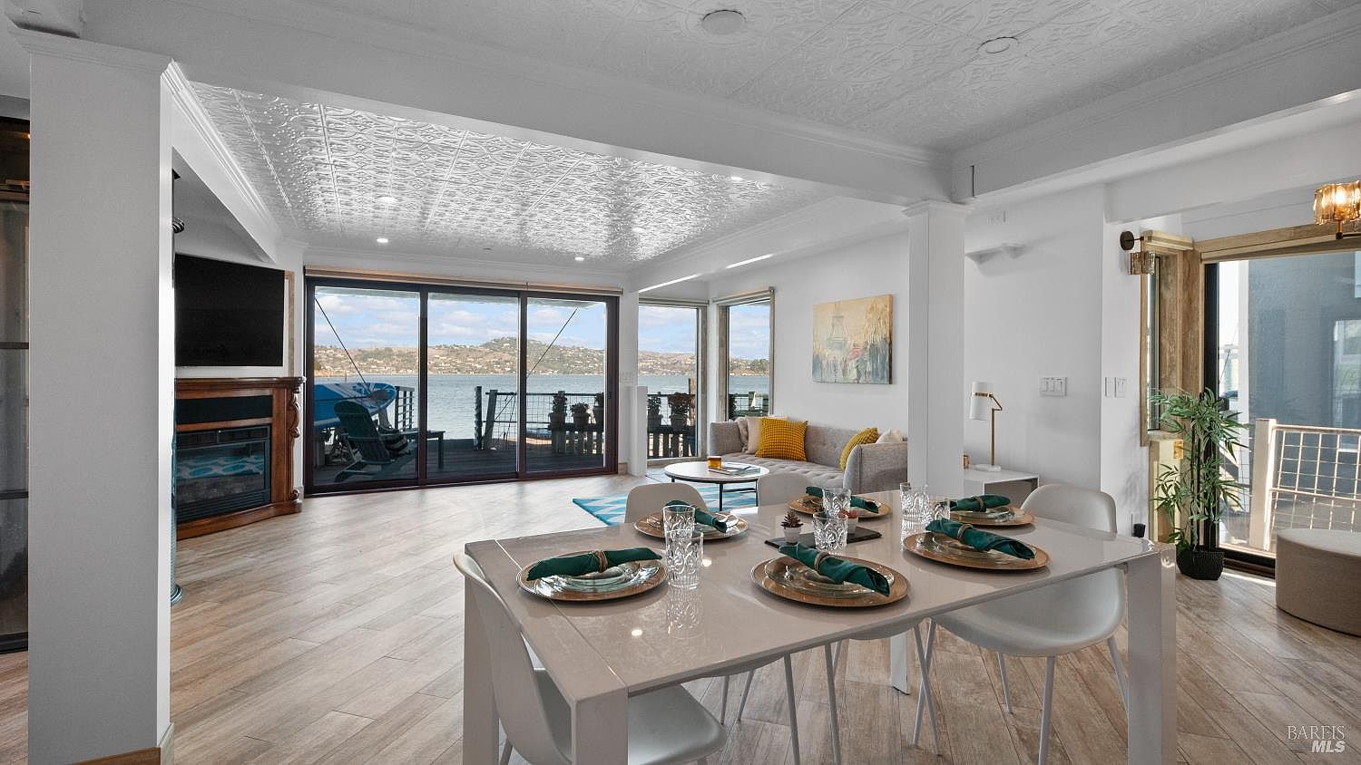 Open-plan dining and living room with dressed dining table, fireplace, and a wall of glass opening onto the deck and bay view.
