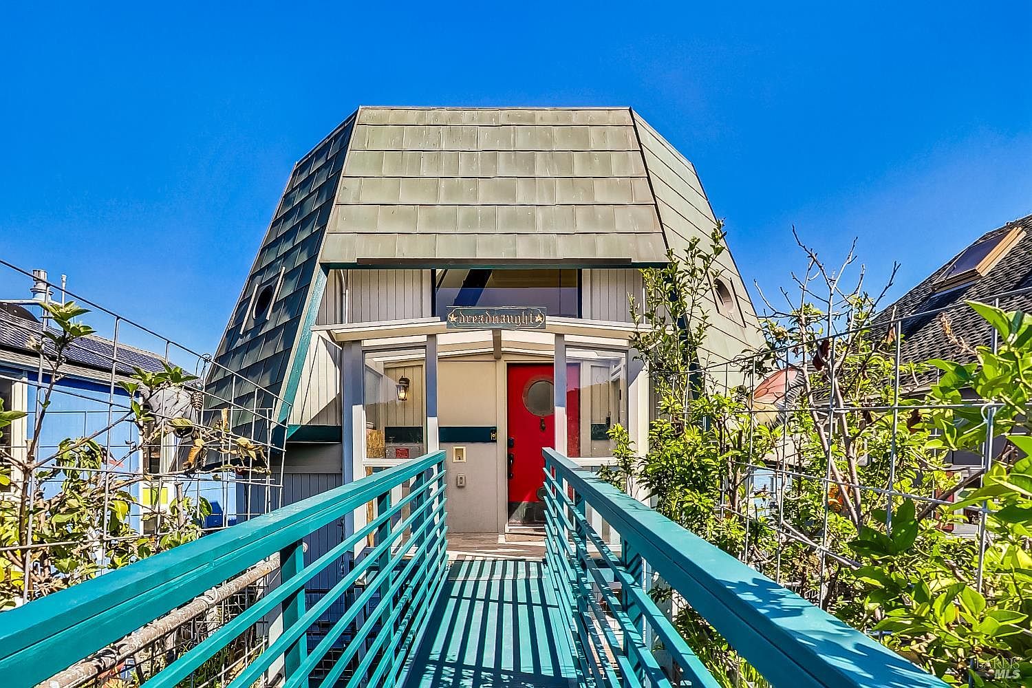 68 Issaquah Dock, the front entry of a custom floating home with a copper mansard roof and red door, approached over a teal-railed bridge under a clear Sausalito sky.